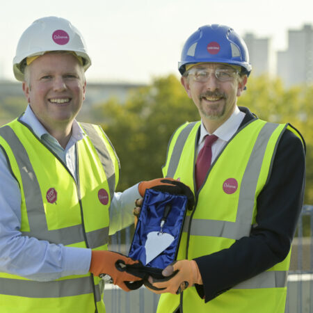 two white men in hi vis clothing and hardhats on top of a construction site building holding a silver plated trowel